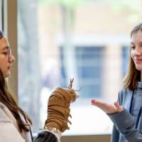 Two middle school girls standing inside in front of a window, one holding a brown hand-puppet of a bug